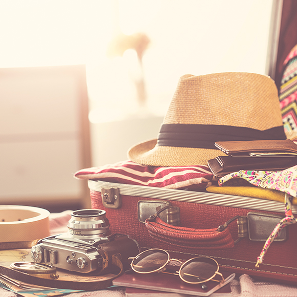 Packed suitcase laying open on bed, complete with a straw hat, camera and sunglasses to promote travel insurance by Evalee Insurance Brokers